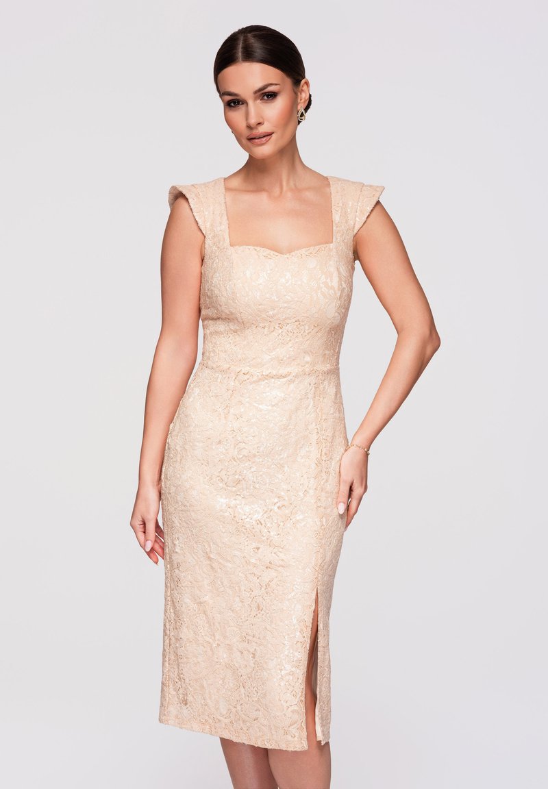 Femme portant une robe sans manches en dentelle beige longueur genou, avec un décolleté carré et une fente latérale, posant devant un fond clair uni.