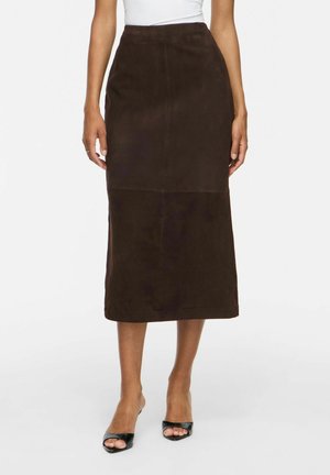 Femme portant une jupe midi en daim marron foncé avec une taille haute ajustée et des talons ouverts noirs, debout devant un fond blanc.
