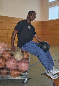 Basketballspiller iført sort t-shirt med logo, denim jeans, siddende på en trådkurv fyldt med basketballer i en sportshal. Basketball i hånden.