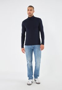 Tricou cu guler înalt, mâneci lungi, de culoare bleumarin, dintr-un material ușor, asortat cu blugi de denim bleu deschis și teniși alb-negru, pe un fundal simplu.