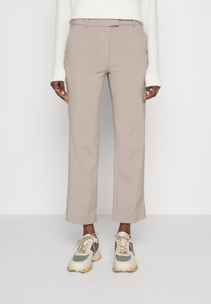 Mannequin portant un pantalon beige tailleur court, un pull blanc et des baskets blanches épaisses avec des détails verts et beiges, debout devant un fond uni.