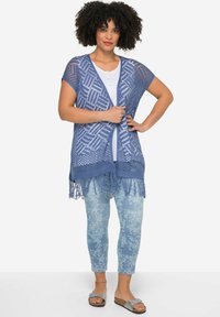 Vest all'uncinetto di colore azzurro chiaro con apertura frontale, motivo geometrico e dettagli con frange, abbinato a jeans leggeri a fantasia e sandali piatti.