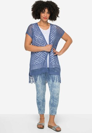 Chaleco de ganchillo azul claro con abertura frontal, patrón geométrico y detalle de flecos, combinado con jeans claros con estampado y sandalias planas.