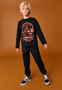 Svart långärmad skjorta med orange grafisk design och text, kombinerad med svarta cargobyxor och svarta sneakers med vita sulor.