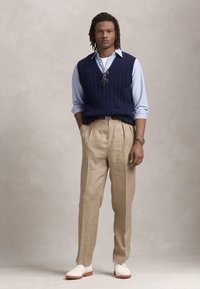 Marineblå kabelstrik vest over en lyseblå skjorte, parret med beige ternede bukser og hvide sko med brun sål. Modellen står lige op.