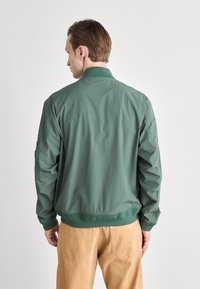 Zelená bomber bunda s ribovaným límcem a manžetami, lehká tkanina, elastický lem, nošená s béžovými kalhotami, s jednoduchým designem.