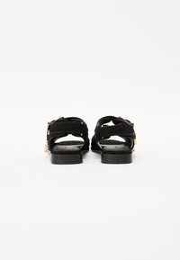 Sorte nubuck sandaler med flate såler, som har to stropper og detaljer i gullfarge. Bakfra, som viser åpen design og teksturert såle.