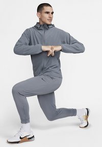 Hall halli spordijakk kapuutsiga ja eest lukuga, sobitatud kitsenevate pükstega. Modell kannab valgeid ja musti tenniseid ning näitab väljaastumise poosi.