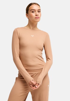 Jeune femme à la peau claire et aux cheveux attachés, portant un haut beige côtelé à manches longues et un pantalon assorti, debout les mains jointes.