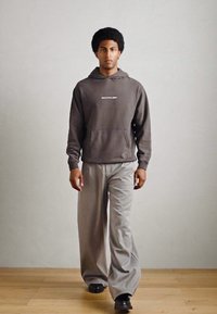 Grijze hoodie met een voorzak en logo op de borst, gecombineerd met grijze wijde broek en zwarte schoenen. Zachte, casual stoftexturen.