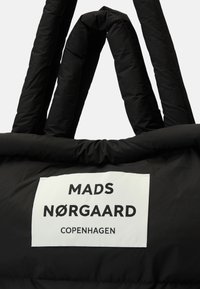 Μαύρη τσάντα tote με μαξιλαρωτό υλικό και παχιές, στρογγυλές λαβές. Φέρει λευκή ετικέτα με την ένδειξη "MADS NØRGAARD COPENHAGEN" εκτυπωμένη με έντονα μαύρα γράμματα.
