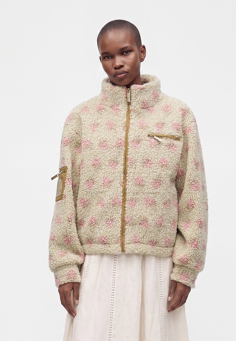 Personne portant une veste polaire beige avec des motifs de cœurs roses, une fermeture éclair devant, ainsi que des poches zippées sur la poitrine et les bras, par-dessus une jupe blanche.