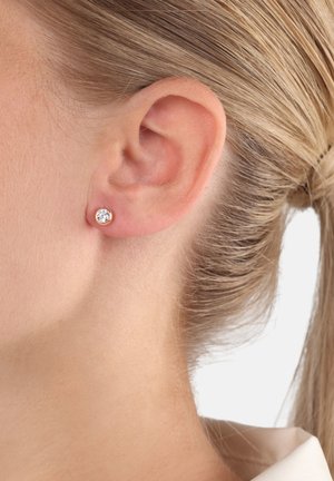Primer plano de la oreja de una persona con cabello rubio que lleva un pequeño pendiente circular con una gema transparente en un engaste dorado.