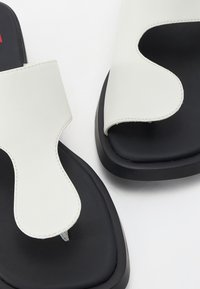 Vita lädersandaler med en bred rem och utskuret mönster, placerade på en svart gummisula. Slät yta med kontrasterande sömdetaljer.