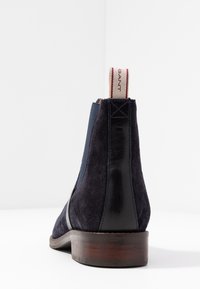 Marineblauwe suède enkelboot met leren accenten, een houten hak en een gestreepte stoffen lus aan de achterkant voor gemakkelijke aan- en uittrekken.