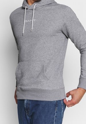 Mies pukeutuneena vaaleanharmaaseen GANT-huppariin, jossa on valkoiset nyörit ja etutasku, yhdistettynä sinisiin farkkuihin.