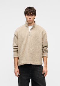 Νεαρός άνδρας που φοράει ένα μπεζ φλις πουλόβερ με μισό φερμουάρ και μαύρα τζιν, στέκεται μπροστά σε λευκό απλό φόντο.