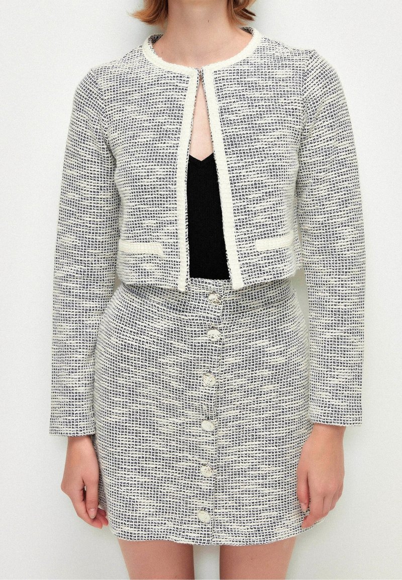 Femme portant une jupe texturée blanche et noire boutonnée et une veste courte assortie par-dessus un haut noir, debout devant un fond uni.