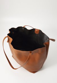 Brun skinn totebag med strukturert ytre, bølgete toppkant og to lange håndtak; innsiden er svart. Minimalistisk design.