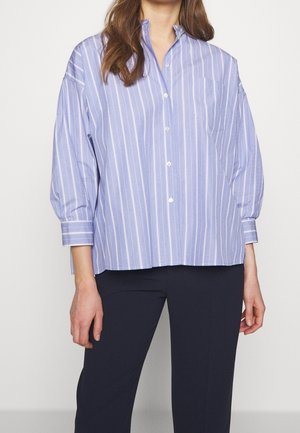 Woman wearing a loose, light blue striped button-up shirt with 3/4 sleeves and dark pants, standing against a plain white background.