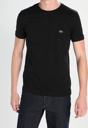 Mand iført en ensfarvet sort kortærmet t-shirt med et lille krokodillelogo på venstre bryst og mørkeblå jeans.