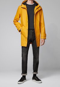 Žlutá voděodolná bunda do deště s kapucí, černými akcenty a patentkami. Nesená přes tmavý svetr a šedé džíny s ohrnutými manžetami. Černé tenisky.