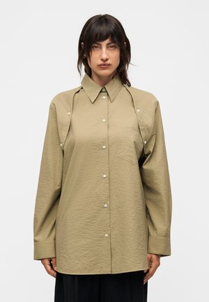 Γυναίκα με σκούρα μαλλιά φοράει υπερμεγέθη μπεζ πουκάμισο με κουμπιά και κοψίματα στους ώμους, στέκεται μπροστά σε απλό φωτεινό φόντο.