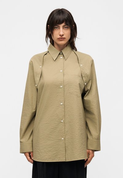 Femme aux cheveux foncés portant une chemise beige surdimensionnée à boutons avec des découpes aux épaules, debout devant un fond uni clair.
