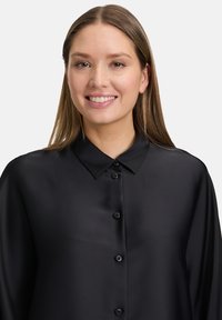 Camicia nera con bottoni realizzata in tessuto liscio, con collo a punte e maniche lunghe. Presenta una leggera lucentezza e sei bottoni neri.