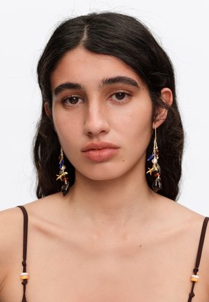 Joven mujer con largo cabello oscuro lleva un top marrón con tirantes finos y pendientes largos colgantes con cuentas y amuletos sobre un fondo blanco.