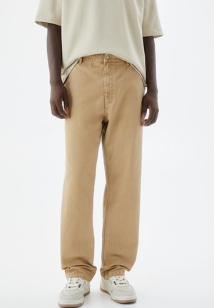 Beige bomulds twill bukser, rummelig pasform, frontlukning med knap og lynlås, diskrete syninger, parret med hvide sneakers, glat tekstur.