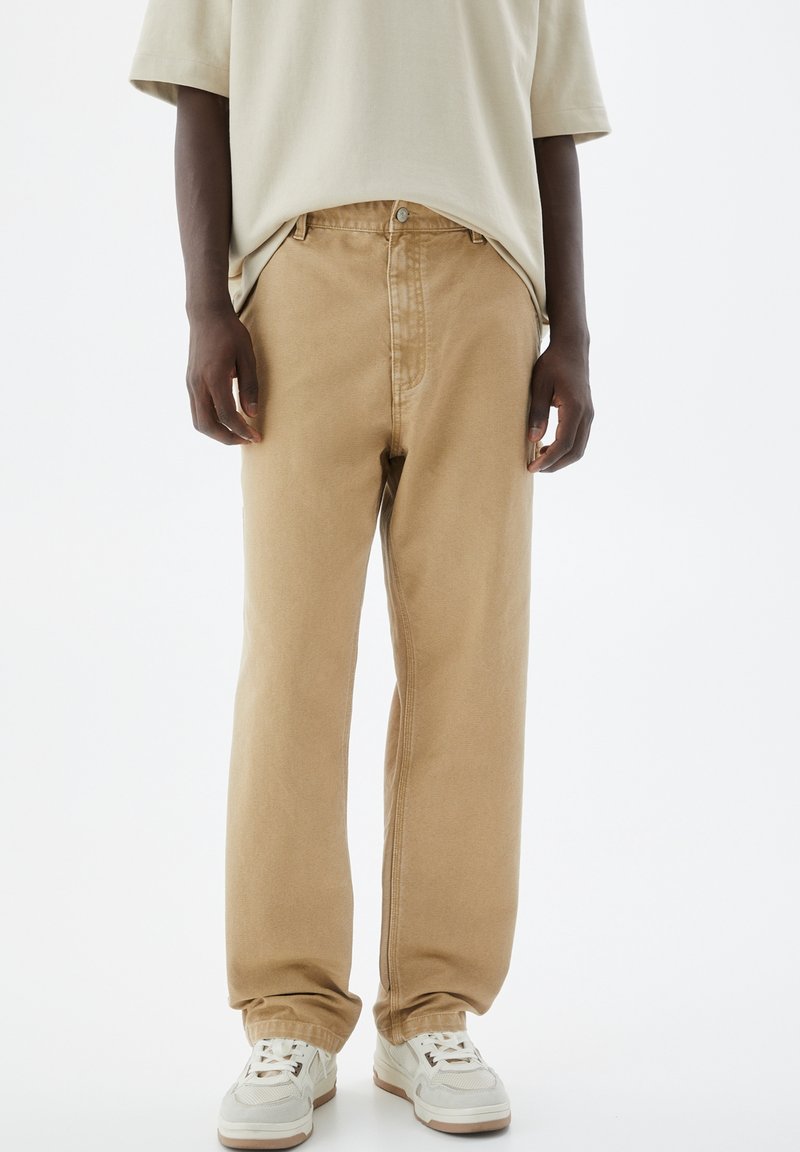 Beiga bomulls twill byxor, rymlig passform, frontknapp och dragkedja, diskret sömnadsdetalj, parat med vita sneakers, slät textur.
