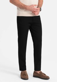 Schwarze Slim-Fit-Hosen aus glattem Stoff. Kombiniert mit braunen Wildleder-Loafers, die mit einem Quasten-Detail versehen sind. Das Modell steht lässig.
