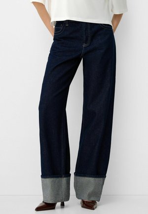 Donkerblauwe wijd uitlopende jeans met grote omgeslagen boorden, gedragen met bruine, puntige, met hakken voorziene schoenen en een wit shirt, model staand met handen in de zakken.