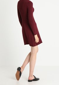 Vestito bordeaux con corsetto a coste e gonne a campana; maniche lunghe; indossato con scarpe nere senza lacci con suole piatte e dettagli argentati.