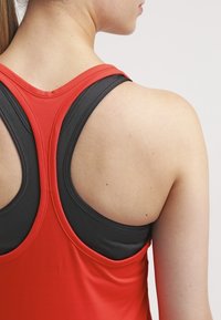 Punainen ja musta urheilutanktoppi, jossa on r Racerback-malli. Kangas on sileä ja joustava, eri värit erottuvat hyvin, ja saumat ovat näkyvissä.