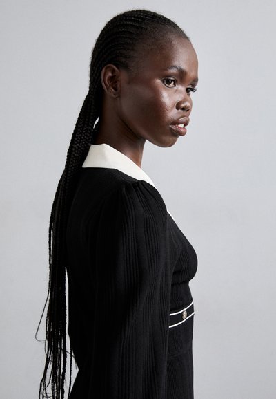 Vestido negro de canalé con un collar blanco y un botón decorativo, con mangas largas y una silueta ajustada, destacado con una franja horizontal.