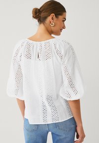 White blouse with puff sleeves, featuring lace cut-out details and a keyhole back. Made from lightweight fabric, paired with denim jeans.