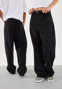 Vidde-ben svarte denimjeans med høy midje, knappelukking og to baklommer; kombinert med mønstrede slip-on sko.