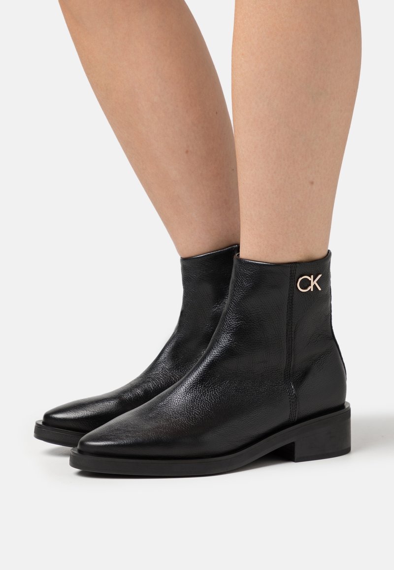Bottines en cuir noir avec un bout pointu, une finition texturée, un petit talon carré et un accent logo "CK" en or sur le côté.