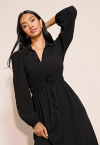 Černé šaty s límcem, dlouhými nafouknutými rukávy a shromážděným pasem se stuhou. Mají strukturovanou tkaninu a splývavý design.