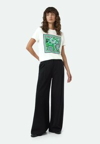 Witte T-shirt met een groene bloemenprint in een vierkant kader; gecombineerd met zwarte wijde broek en zwarte schoenen. Glad stoftextuur.