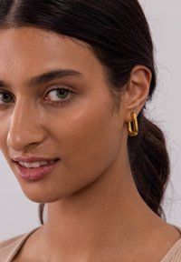 Goldene Kreole mit einer glatten, abgerundeten Form. Die Ohrringe haben eine polierte Oberfläche und ein leicht verlängertes Design, das ihre visuelle Anziehungskraft erhöht.