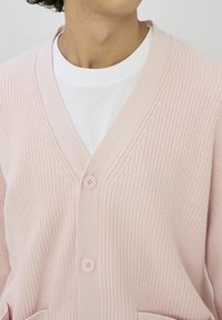 Personne portant un cardigan en tricot rose clair boutonné par-dessus un t-shirt blanc à col rond, coupé au niveau du cou et de la poitrine.