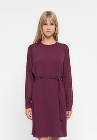 Rochie cu mânecă lungă de culoare mov închis, cu un corp texturat și mâneci din material transparent cu model în zigzag. Talie legată pentru ajustarea potrivirii.