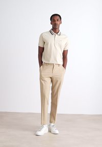 Beige polo shirt met een zwart-wit gestreepte kraag, gecombineerd met lichtbruine broeken en witte sneakers. Gladde stof, slim fit.