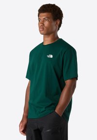 Ung mand med kort krøllet hår, iført en grøn The North Face t-shirt og sorte bukser, stående med den ene hånd i lommen.