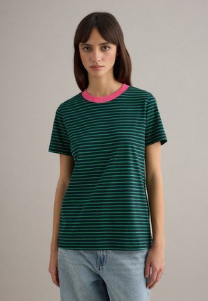 Vrouw draagt een donker groen en marineblauw gestreept T-shirt met een roze kraag en lichtblauwe jeans, staand tegen een effen achtergrond.