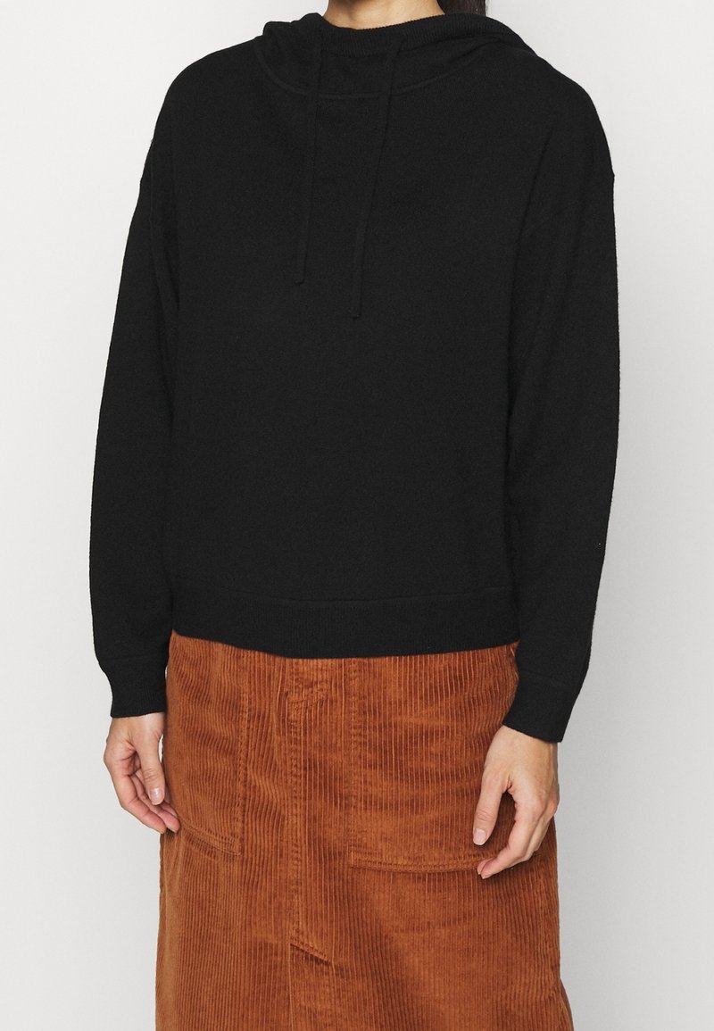Femme portant un sweat à capuche noir à manches longues et une jupe en velours côtelé marron taille haute, debout devant un fond uni.