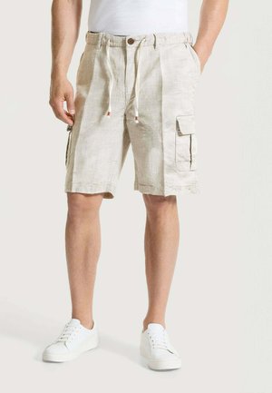 Uomo con pantaloncini cargo beige chiaro con coulisse e bottone, abbinati a sneaker bianche e t-shirt bianca, in piedi davanti a uno sfondo semplice.